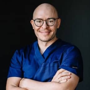 Porträt von Dr. Adorján Szakál, Mundchirurg bei CompletDent Budapest — in dunkelblauer Praxiskleidung vor dunklem Hintergrund